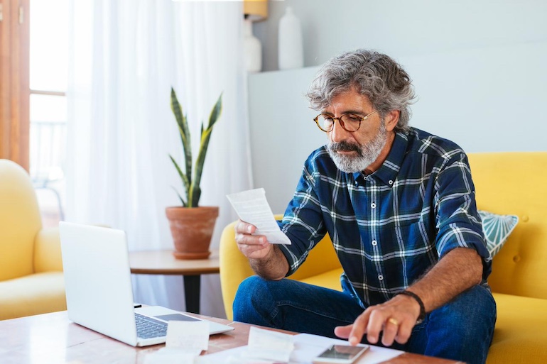 Älterer Mann sitzt am Laptop, auf dem Tisch liegt ein Taschenrechner, auf welchen er tippt, während er auf ein Dokument blickt