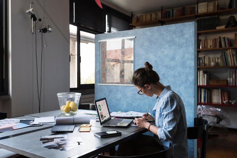 Maklerin mit Brille und hochgebundenen Haaren sitzt an einem Schreibtisch mit vielen Dokumenten, Laptop und Notizen und arbeitet eine Checkliste ab